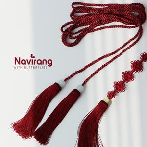 May include: A deep red decorative cord with tassels and a knotted design. The cord features two tassels with white beads and a series of square knots. The logo "Navirang with Butterflies" is visible in the upper left corner.