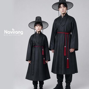 May include: Two individuals wearing black traditional Korean hanbok robes with long sleeves, high collars, and red waist ties featuring hanging tassels. Both wear wide-brimmed black hats with chin straps. The text Navirang WITH BUTTERFLIES is visible.