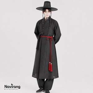 May include: A black traditional Korean hanbok robe featuring long sleeves, a wrap-style front, and a vibrant red cord belt with tassels. The outfit includes a wide-brimmed black hat and a long beaded necklace. The text Navirang WITH BUTTERFLIES is visible.