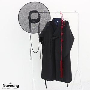 May include: A black wide-brimmed mesh hat with a beaded chin strap, a black long-sleeved robe with a wrap-front closure, and a long red decorative cord with intricate knots and a tassel, all hanging on a white rack. Text reads Navirang WITH BUTTERFLIES.