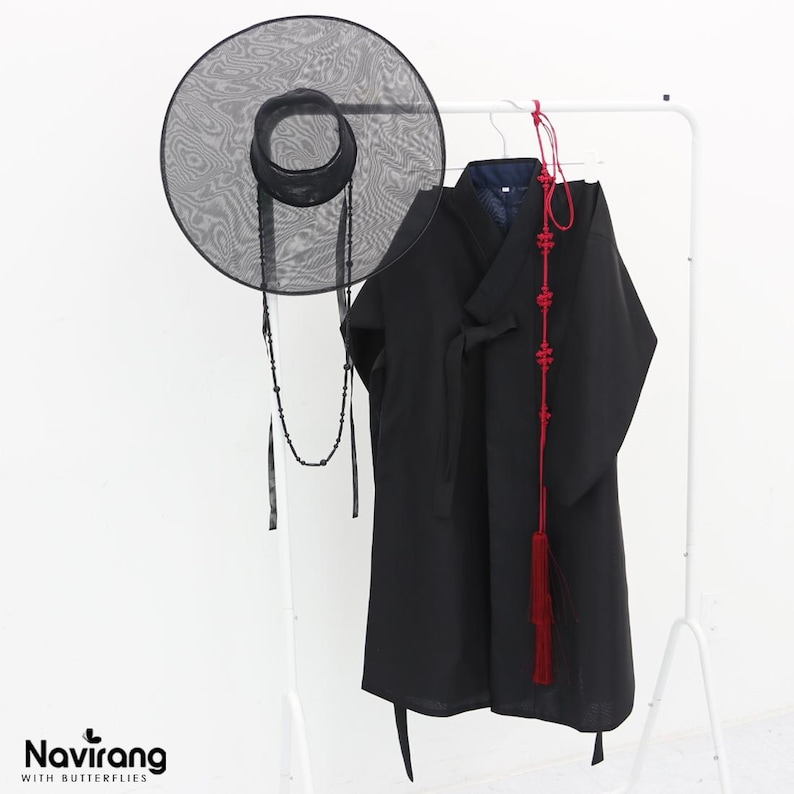 May include: A black wide-brimmed mesh hat with a beaded chin strap, a black long-sleeved robe with a wrap-front closure, and a long red decorative cord with intricate knots and a tassel, all hanging on a white rack. Text reads Navirang WITH BUTTERFLIES.