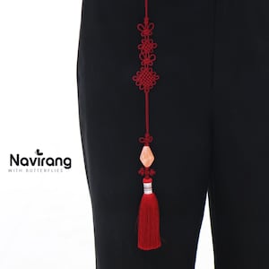 Può includere: Un nappa decorativa rossa con una serie di disegni annodati e una gemma color pesca. La nappa è appesa su uno sfondo nero. Il testo "Navirang with Butterflies" si trova nell'angolo in basso a sinistra.