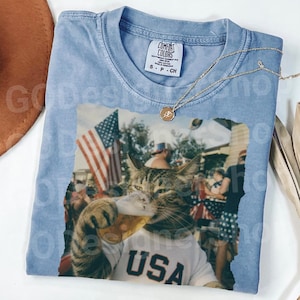 May include: Light blue t-shirt with a graphic of a cat drinking beer at a patriotic event. The cat wears a white shirt with "USA" printed on it. A gold necklace and a Comfort Colors tag are visible.
