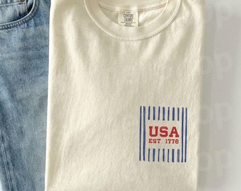 Puede incluir: Una camiseta color crema con un gráfico de USA y unos vaqueros azules. La camiseta presenta un diseño rectangular con líneas azules verticales, la palabra "USA" en rojo y "EST. 1776". Los vaqueros son de color azul claro.