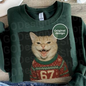 Green sweatshirt with a laughing cat wearing a festive red and green sweater featuring the number 67 in a humorous Christmas design.