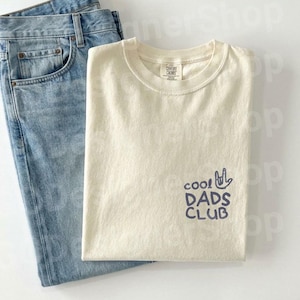 May include: A cream-colored t-shirt with the text "cool DADS CLUB" in navy blue, paired with light blue denim jeans. The t-shirt has a crew neck and a small logo on the upper back.