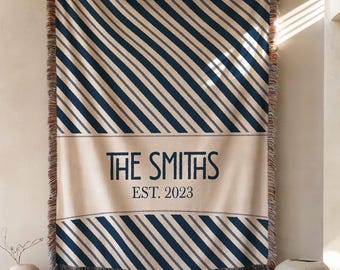 May include: A beige throw blanket with navy and gray diagonal stripes. The blanket features a rectangular section with the text "THE SMITHS EST. 2023" in navy. The edges are fringed.