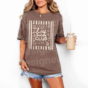 May include: A brown t-shirt with the words "boy mom CLUB" in white, along with various illustrations. The shirt has a boxy fit and short sleeves. The text "est. 2026" is also included. The shirt is worn with denim shorts.