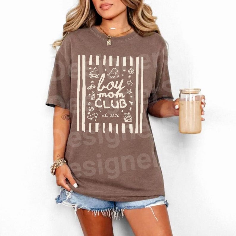 May include: A brown t-shirt with the words "boy mom CLUB" in white, along with various illustrations. The shirt has a boxy fit and short sleeves. The text "est. 2026" is also included. The shirt is worn with denim shorts.