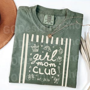 May include: Olive green t-shirt with the words "girl mom CLUB est. 2026" in a white graphic. The design includes a crown, rainbow, unicorn, and other small illustrations. A gold necklace rests on the shirt.