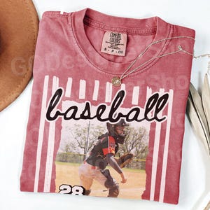 May include: A maroon baseball t-shirt with the word "baseball" in a script font. The shirt features a graphic of a baseball player and the number 28. A silver necklace with a pendant is draped across the shirt.