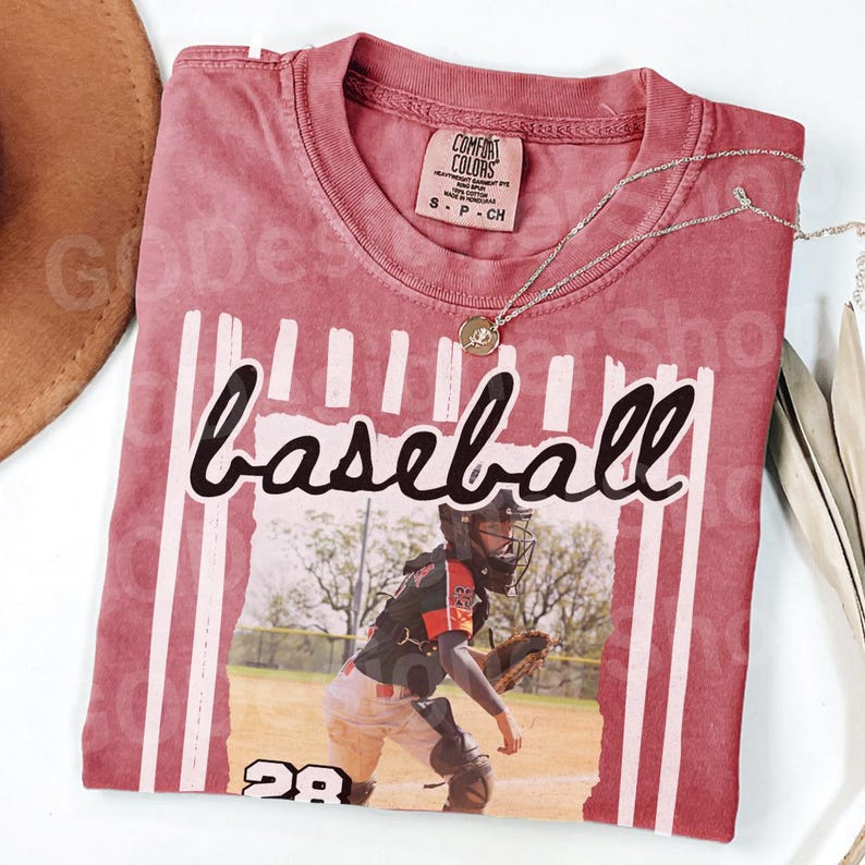 May include: A maroon baseball t-shirt with the word "baseball" in a script font. The shirt features a graphic of a baseball player and the number 28. A silver necklace with a pendant is draped across the shirt.