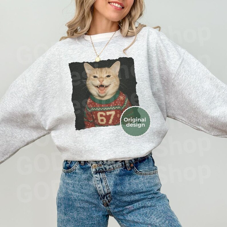 May include: A light gray sweatshirt featuring a cat wearing a Christmas sweater with the number 67. The cat image is set against a black square, with a green circle that says "Original design". The model is wearing blue jeans.