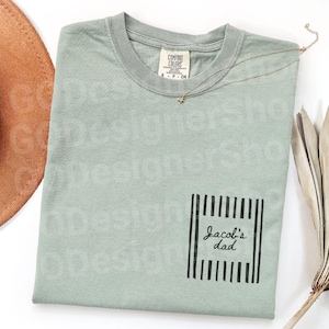 May include: A sage green t-shirt with a pocket featuring the text "Jacob's dad" in a black, vertical striped frame. A gold star necklace and a brown hat are visible. The shirt is made by Comfort Colors.