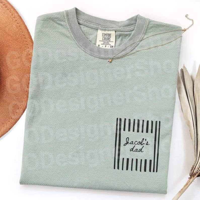 May include: A sage green t-shirt with a pocket featuring the text "Jacob's dad" in a black, vertical striped frame. A gold star necklace and a brown hat are visible. The shirt is made by Comfort Colors.