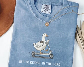 May include: A blue Comfort Colors t-shirt featuring a cartoon goose riding a scooter. The goose is holding a book and the text "OFF TO REJOICE IN THE LORD" is printed below the illustration. A gold necklace is draped across the shirt.