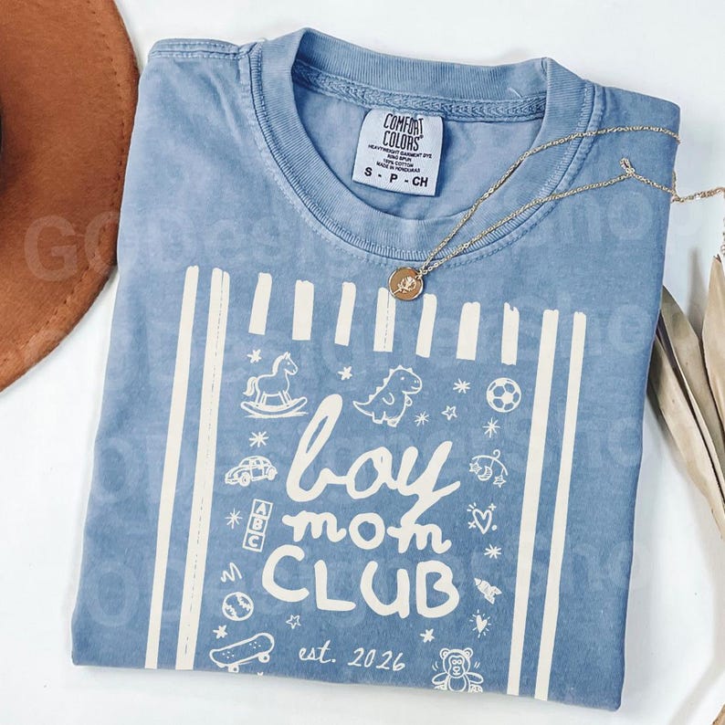 May include: A light blue Comfort Colors t-shirt with the words "boy mom CLUB" and the year "est. 2026" printed in white. The shirt features white vertical stripes and various small illustrations of toys. A gold necklace is draped across the shirt.