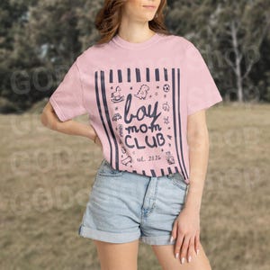 May include: A light pink t-shirt with the text "boy mom CLUB" and the date "est. 2026" in a navy blue font. The shirt features a navy blue striped design and small illustrations of toys.