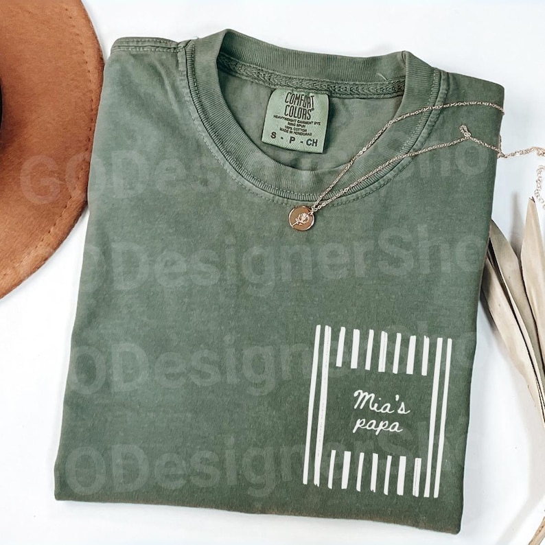 May include: An olive green t-shirt with the text "Mia's papa" in a white rectangular design. A gold necklace with a pendant rests on the shirt. The shirt is a Comfort Colors brand.