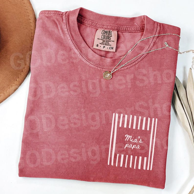 May include: A red Comfort Colors t-shirt with a pocket design that reads "Mia's papa" in white. A silver necklace with a pendant is draped across the shirt. A brown hat and dried leaves are in the background.