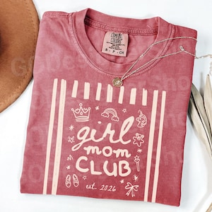 May include: A red Comfort Colors t-shirt with the words "girl mom CLUB" and the date "est. 2026" printed in a cream color. The shirt features a crown, rainbow, unicorn, and other graphics. A silver necklace with a pendant is draped across the shirt.
