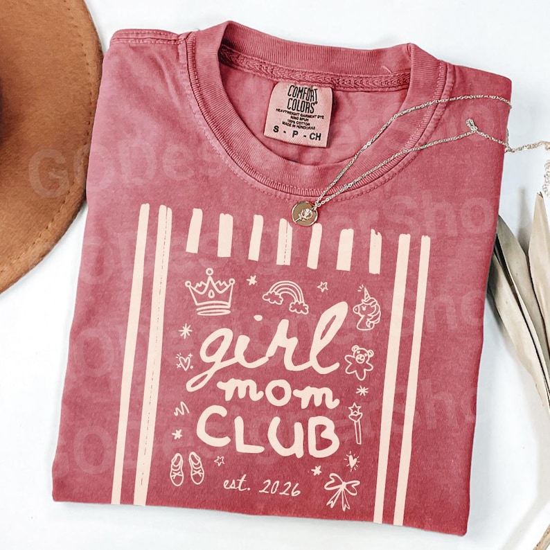 May include: A red Comfort Colors t-shirt with the words "girl mom CLUB" and the date "est. 2026" printed in a cream color. The shirt features a crown, rainbow, unicorn, and other graphics. A silver necklace with a pendant is draped across the shirt.