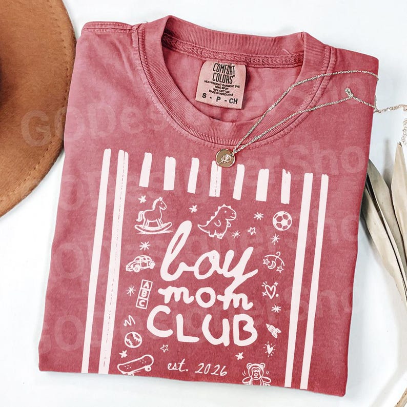 May include: A red t-shirt with the words "boy mom CLUB" and the text "est. 2026" in white. The shirt has a crew neck and short sleeves. A silver necklace with a pendant is draped across the shirt.