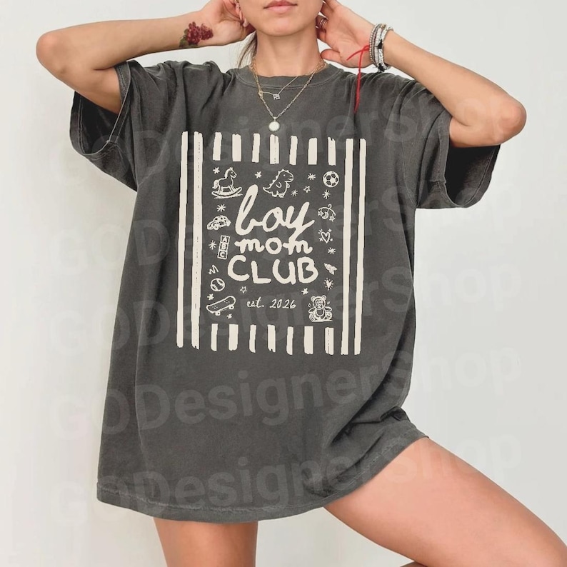May include: Dark gray t-shirt with a white graphic that reads "boy mom CLUB est. 2026." The graphic is framed by vertical stripes and includes illustrations of toys. The shirt has short sleeves and a relaxed fit.