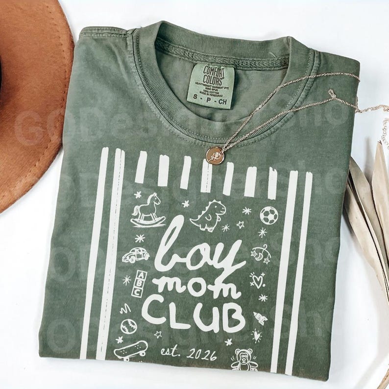 May include: An olive green t-shirt with the words "boy mom CLUB est. 2026" in white, along with various illustrations. A gold necklace rests on the shirt. The shirt is folded on a white surface.