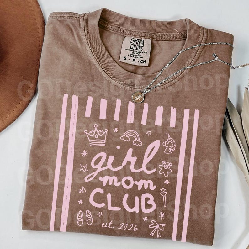 May include: Brown Comfort Colors t-shirt with a pink graphic that says "girl mom CLUB est. 2026". The design includes a crown, rainbow, unicorn, and other illustrations. A silver necklace with a pendant is on the shirt.