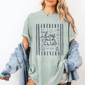 May include: A light green t-shirt with a navy blue graphic that reads "boy mom CLUB est. 2026". The graphic is framed by vertical lines and includes illustrations of a rocking horse, dinosaur, and other toys. The shirt is worn with a denim jacket.