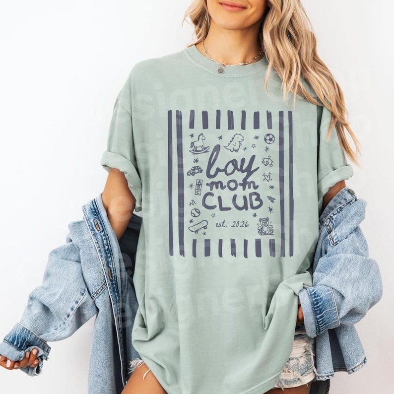May include: A light green t-shirt with a navy blue graphic that reads "boy mom CLUB est. 2026". The graphic is framed by vertical lines and includes illustrations of a rocking horse, dinosaur, and other toys. The shirt is worn with a denim jacket.