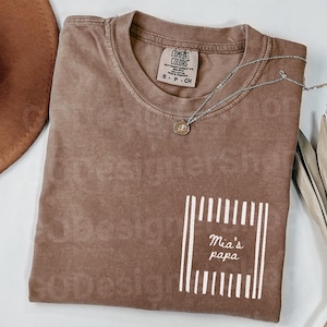May include: A brown Comfort Colors t-shirt with the text "Mia's papa" in a white rectangular design. A silver necklace with a small pendant is draped across the shirt. The shirt is folded neatly.