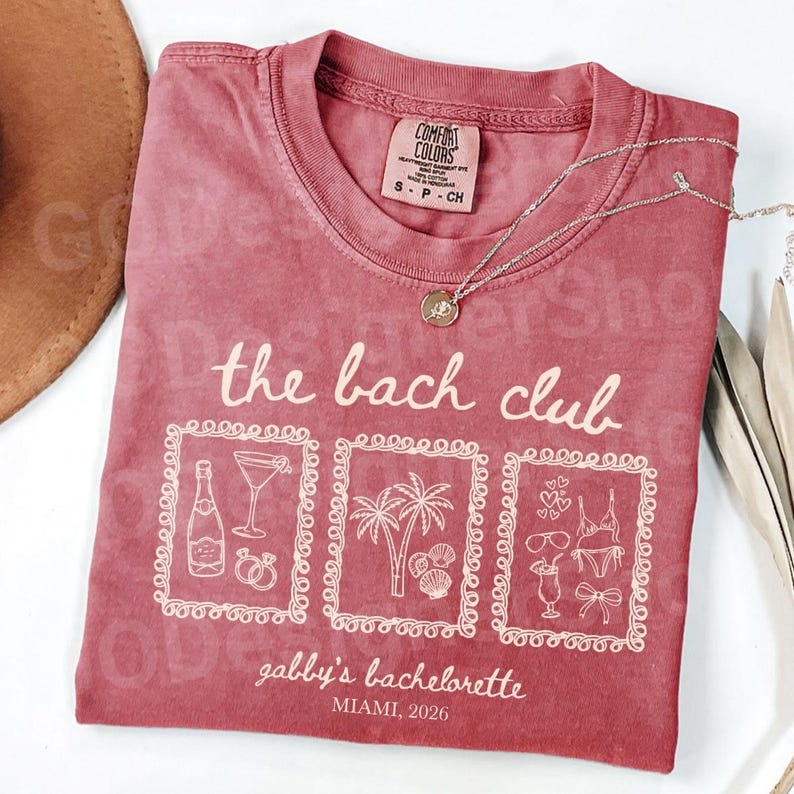 May include: A red Comfort Colors t-shirt with the text "the bach club" and three framed illustrations. The illustrations include a champagne bottle, martini glass, palm trees, seashells, and swimwear. A silver necklace with a pendant is on the shirt.