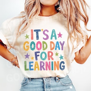 May include: A cream-colored short-sleeve t-shirt featuring the multi-colored text IT'S A GOOD DAY FOR LEARNING in a distressed, block-letter style. The design includes small colorful stars interspersed among the words in shades of red, blue, green, yellow, and purple.
