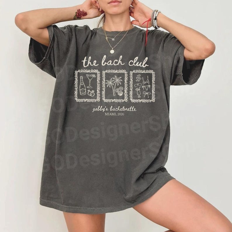 May include: Dark gray t-shirt with white text and graphics. The shirt reads "the bach club" above three framed illustrations of a bottle, cocktail, palm trees, and flowers. Below the illustrations is "gabby's bachelorette, Miami 2026".