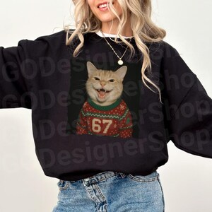 May include: Black sweatshirt featuring a smiling orange tabby cat wearing a red and green Christmas sweater with the number 67. The cat's mouth is open, showing its teeth. The sweatshirt is a casual style.