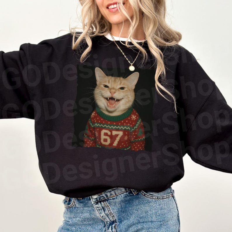 May include: Black sweatshirt featuring a smiling orange tabby cat wearing a red and green Christmas sweater with the number 67. The cat's mouth is open, showing its teeth. The sweatshirt is a casual style.