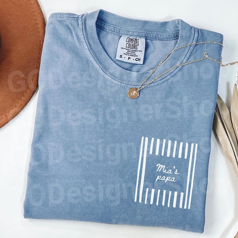 May include: A blue Comfort Colors t-shirt with a pocket design that reads "Mia's papa" in white. A gold necklace with a pendant rests on the shirt. The shirt is size small.
