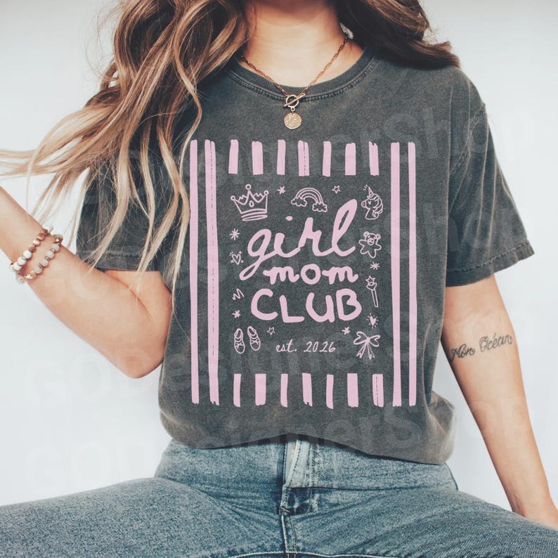 May include: Dark gray t-shirt with a pink graphic that reads "girl mom CLUB est. 2026." The design includes a crown, rainbow, unicorn, and other cute illustrations. The shirt is a casual style.