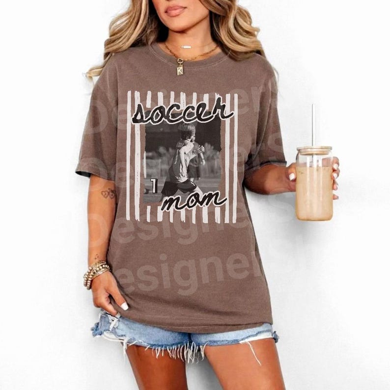 Puede incluir: Camiseta marr&oacute;n con una imagen en blanco y negro de un jugador de f&uacute;tbol y las palabras "soccer mom" en una fuente manuscrita. La camiseta tiene un corte holgado y mangas cortas.
