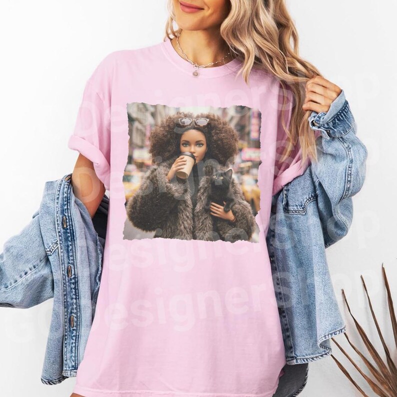 以下が含まれることがあります： ピンク色のTシャツ。大きなアフロヘア、サングラス、毛皮のコートを着た女性が、コーヒーカップと黒猫を持っているグラフィックが特徴です。背景はぼやけた都市です。