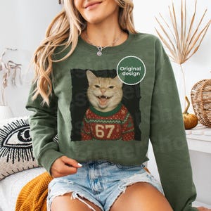 May include: Olive green sweatshirt featuring a smiling cat wearing a red and green Christmas sweater with the number 67. The cat image is framed in black, with a white "Original design" sticker.