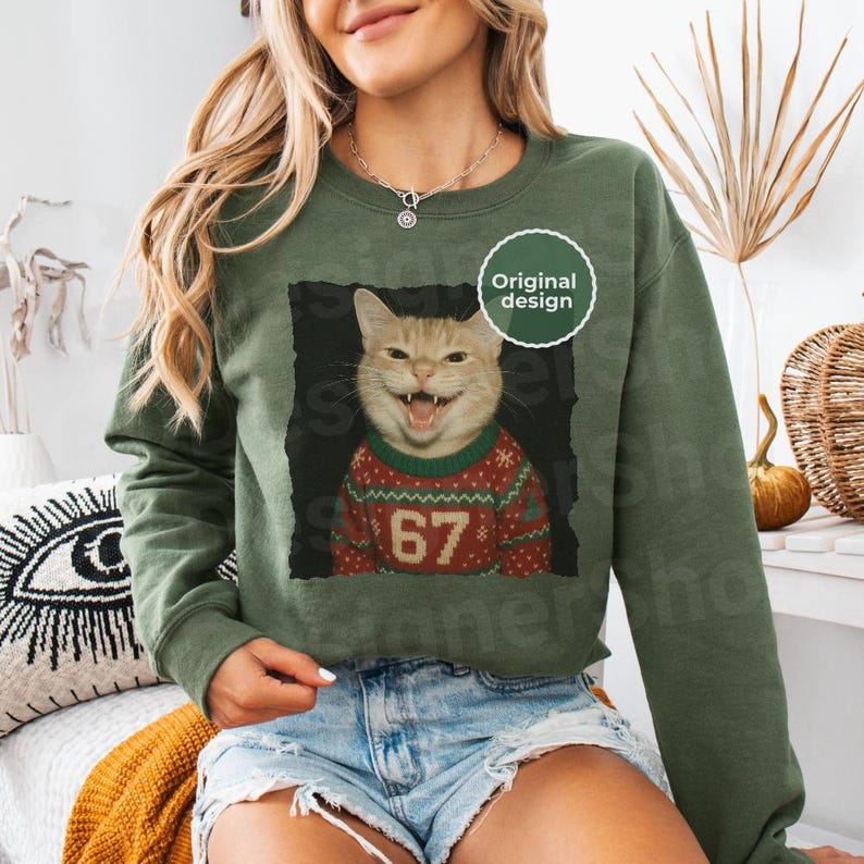 May include: Olive green sweatshirt featuring a smiling cat wearing a red and green Christmas sweater with the number 67. The cat image is framed in black, with a white "Original design" sticker.