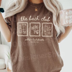 May include: A brown t-shirt with the text "the bach club" and three framed illustrations. The illustrations include a champagne bottle, cocktail, palm trees, seashells, and swimwear. The text "gabby's bachelorette, Miami, 2026" is also printed on the shirt.
