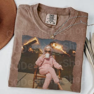 以下が含まれることがあります： サイズSのComfort Colorsラベルが付いた茶色のクルーネックTシャツ。グラフィックプリントには、ピンクのふわふわした服、ヘッドスカーフ、サングラスを身に着け、白いマグカップを持って椅子に座っている人形が、炎を背景に描かれています。