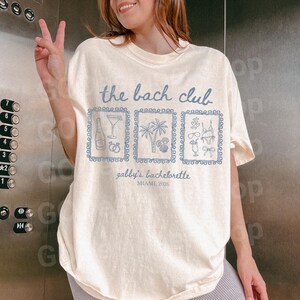 May include: Cream-colored t-shirt with the text "the bach club" in light blue. Below the text are three framed illustrations of a cocktail, palm trees, and swimwear. The text "gabby's bachelorette, MIAMI, 2026" is printed below the illustrations.