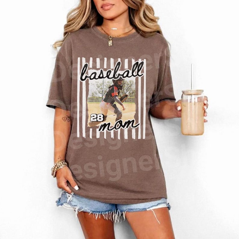May include: Brown t-shirt with the words "baseball mom" in a vertical striped frame. The shirt features a photo of a baseball player and the number 28. The shirt is worn by a person.