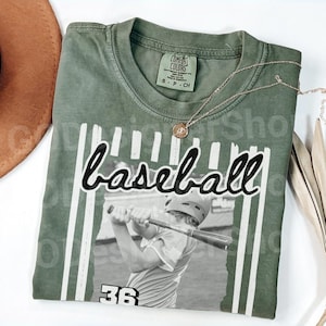 May include: An olive green t-shirt with the word "baseball" in a script font. The shirt features a black and white image of a baseball player swinging a bat, with the number 36 below the image. A gold necklace is draped across the shirt.