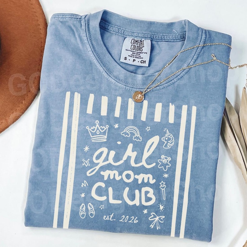 May include: A light blue Comfort Colors t-shirt with the words "girl mom CLUB" and the date "est. 2026" in white. The shirt features a crown, rainbow, unicorn, and other graphics. A gold necklace with a pendant is draped across the shirt.